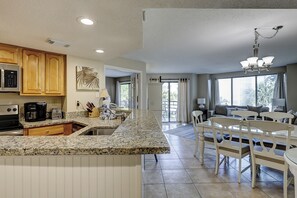 Una cocina con encimeras de granito, armarios de madera, microondas integrado y un comedor con sillas blancas y una mesa.