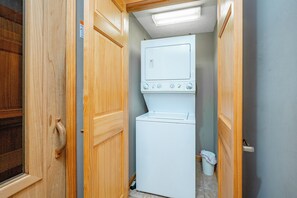 A compact laundry room with a white washer and dryer, wooden doors, and a small trash bin. - Tranquil Lakefront w/ Twin Hot Tubs & Dock (Oakland)