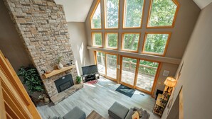 A spacious living area with a stone fireplace, a flat-screen TV, and a large window overlooking a deck. - Tranquil Lakefront w/ Twin Hot Tubs & Dock (Oakland)