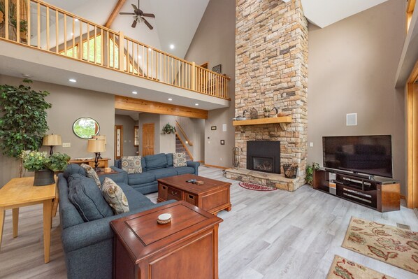 A spacious living room with a stone fireplace, a large TV, and a high ceiling with a wooden balcony.