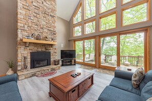 A spacious living room with a stone fireplace, a flat-screen TV, and large windows overlooking a deck.