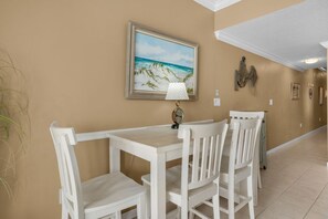 A dining area with a white table and chairs, a framed painting of a beach scene, and a wall-mounted lamp.