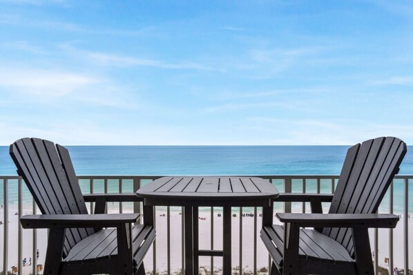 A deck with two Adirondack chairs and a small table, overlooking a beach with clear blue water.
