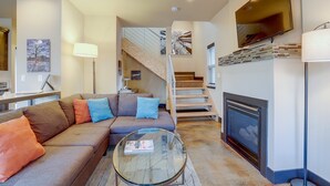Una sala de estar con un sofá modular, una mesa de centro de vidrio, una chimenea, un televisor montado en la pared y una escalera que sube a un nivel superior.