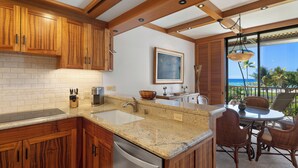 A kitchen with wooden cabinets, granite countertops, and a dishwasher. There is a dining area with wicker chairs and a view of the ocean.