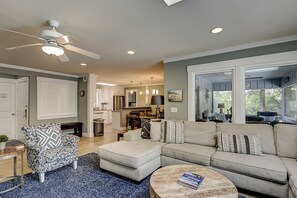 Una sala de estar moderna con sofá, reposapiés y un sillón estampado, un comedor con candelabro, y una cocina con armarios blancos.