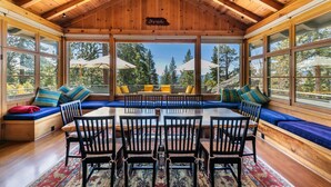 A wooden interior with a large dining table surrounded by chairs, a blue cushioned bench, and a view of trees through large windows.