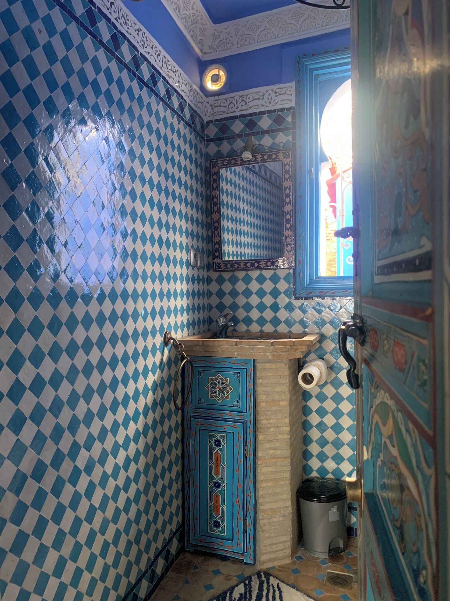 A bathroom with blue and white checkered tiles, a sink with a decorative cabinet, a mirror, and a window with a view.