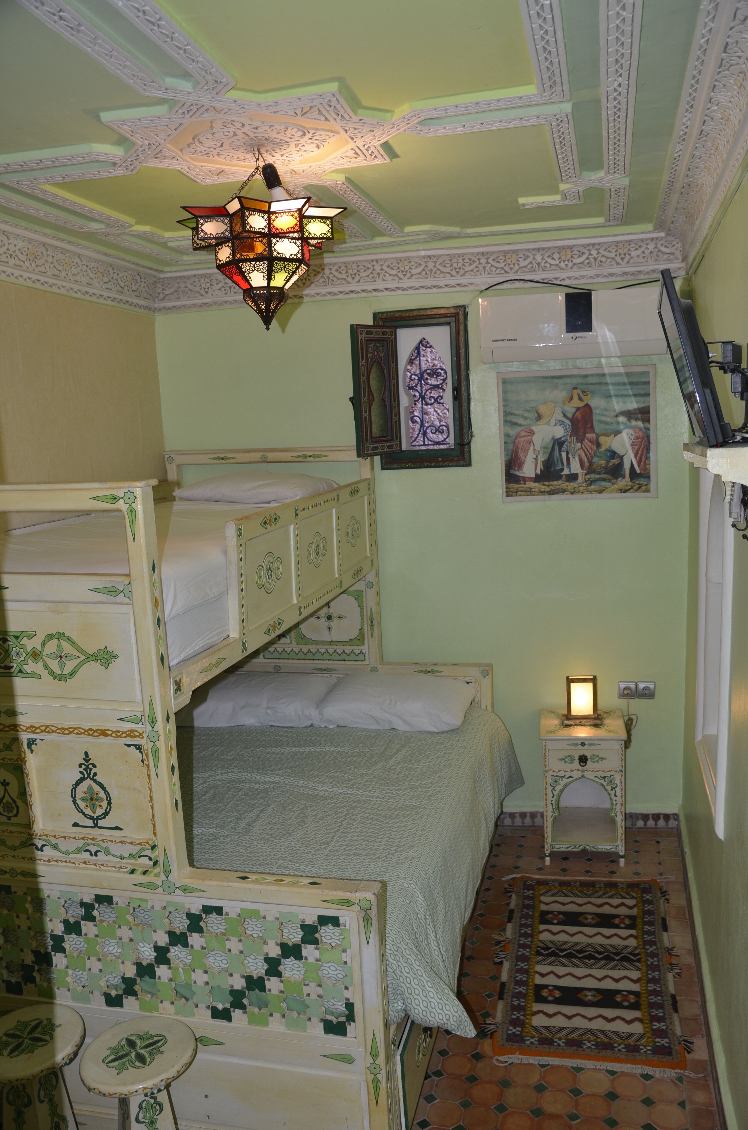 A room with bunk beds, a hanging light fixture, a small bedside table with a lamp, a wall-mounted air conditioner, and a patterned rug.
