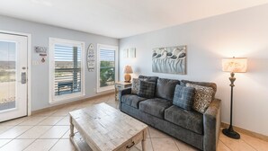 Un salon avec un canapé gris, une table basse en bois, un lampadaire et une vue par une fenêtre équipée de stores.