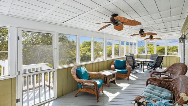Un espacio al aire libre cubierto con muebles de mimbre, ventiladores de techo y grandes ventanales con vistas al entorno.