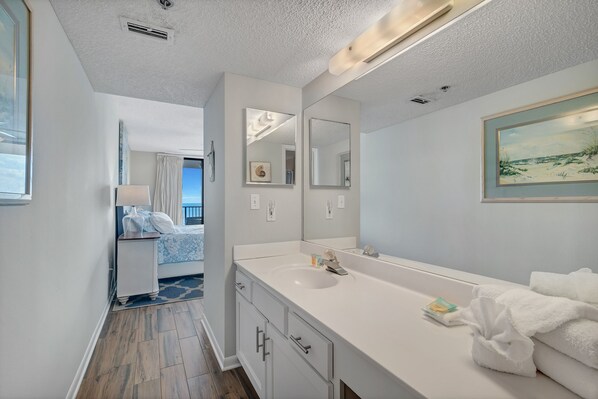 Un baño moderno con un espejo grande, un lavabo blanco y una cómoda. Hay una ventana con vista a la playa y una puerta que da a un dormitorio.