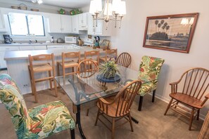 A dining area with a glass table surrounded by wooden chairs, a kitchen in the background, and a framed landscape painting on the wall. - Dune I Townhomes #12 - Beachside Pool, Private Beach Boardwalk, Dune Allen Beach (Santa Rosa Beach)