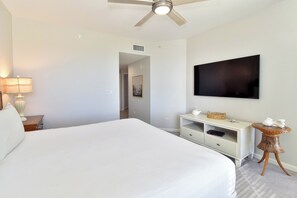 A hotel room with a large bed, a flat-screen TV mounted on the wall, a bedside table with a lamp, and a ceiling fan. - Tile floors, spacious beachfront condo w/private balcony! (Destin)
