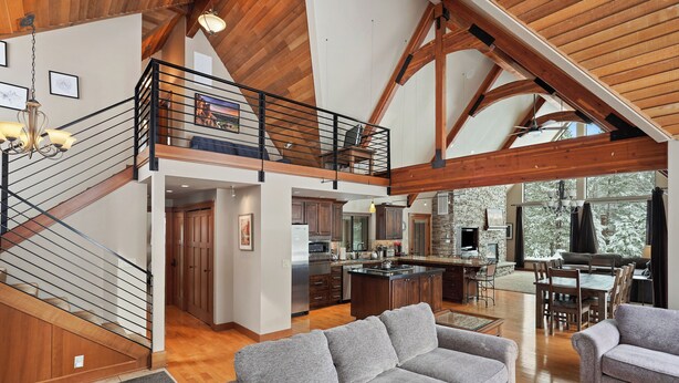 A spacious living area with a high ceiling, wooden beams, a staircase, and a kitchen with a large island.