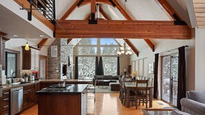 A spacious kitchen with a central island, wooden beams, and large windows offering a view of snow-covered trees.