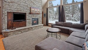 A living room with a stone fireplace, a flat-screen TV, a sectional sofa, and large windows with a snowy view.