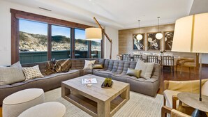 A modern living room with a large sectional sofa, a wooden coffee table, and a dining area with a high table and chairs. Large windows offer a view of the outdoors. - New Upgrades | One Steamboat Place 703 Sugar Loaf (Steamboat Springs)