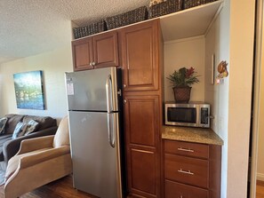 A compact kitchen with a stainless steel refrigerator, wooden cabinets, a microwave, and a potted plant.