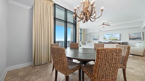 A dining area with a round table, wicker chairs, a chandelier, and large windows with curtains. - Oasis, Formerly Phoenix West II, Beach front, Lazy River/Water Slide!! (Orange Beach)