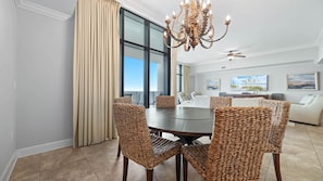 A dining area with a round table, wicker chairs, a chandelier, and large windows with curtains.