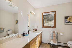 A modern bathroom with a large mirror, two sinks, a window with a view of a forest, and a towel rack with multiple towels. - Mountain Dream: 4 br, fireplace, a/c, game room, deck, hot tub, views! (Stowe)