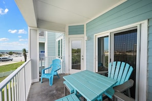 A balcony with a table and chairs, a sliding glass door, and a view of the beach and parking lot.