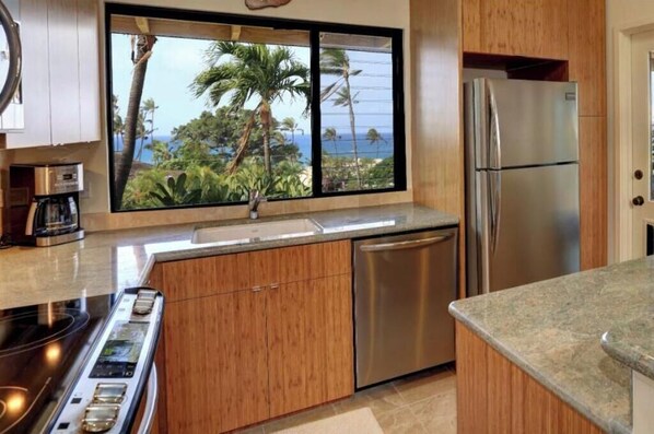 A modern kitchen with wooden cabinets, a stainless steel refrigerator, a dishwasher, and a window with a view of palm trees and the ocean.