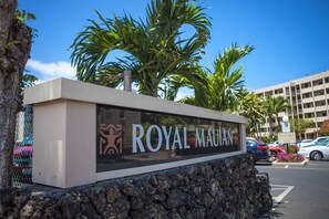 A sign for 'ROYAL MAUIAN' with a stone base, palm trees, and a parking lot in the background. - Experience Oceanfront Grandeur: Royal Mauian #418 - Spectacular Views Await! (Kihei, Maui)