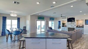 A modern kitchen with a large island, wooden flooring, and a dining area with blue chairs. - Front Row | Pet-Friendly, Stunning Beach Views (Galveston)
