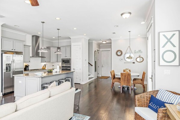 A modern kitchen with white cabinetry, stainless steel appliances, and pendant lighting. There is a dining area with wicker furniture and a white table.