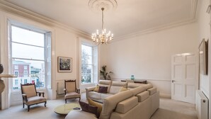A spacious living room with a large beige sectional sofa, two armchairs, a round coffee table, and a chandelier. There are large windows providing natural light and a view of buildings outside. - The Patrick Kavanagh Suite - hiphipstay (Dublin)