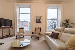 A living room with a sofa, two armchairs, a coffee table, and a television. There are large windows with a view of buildings outside.