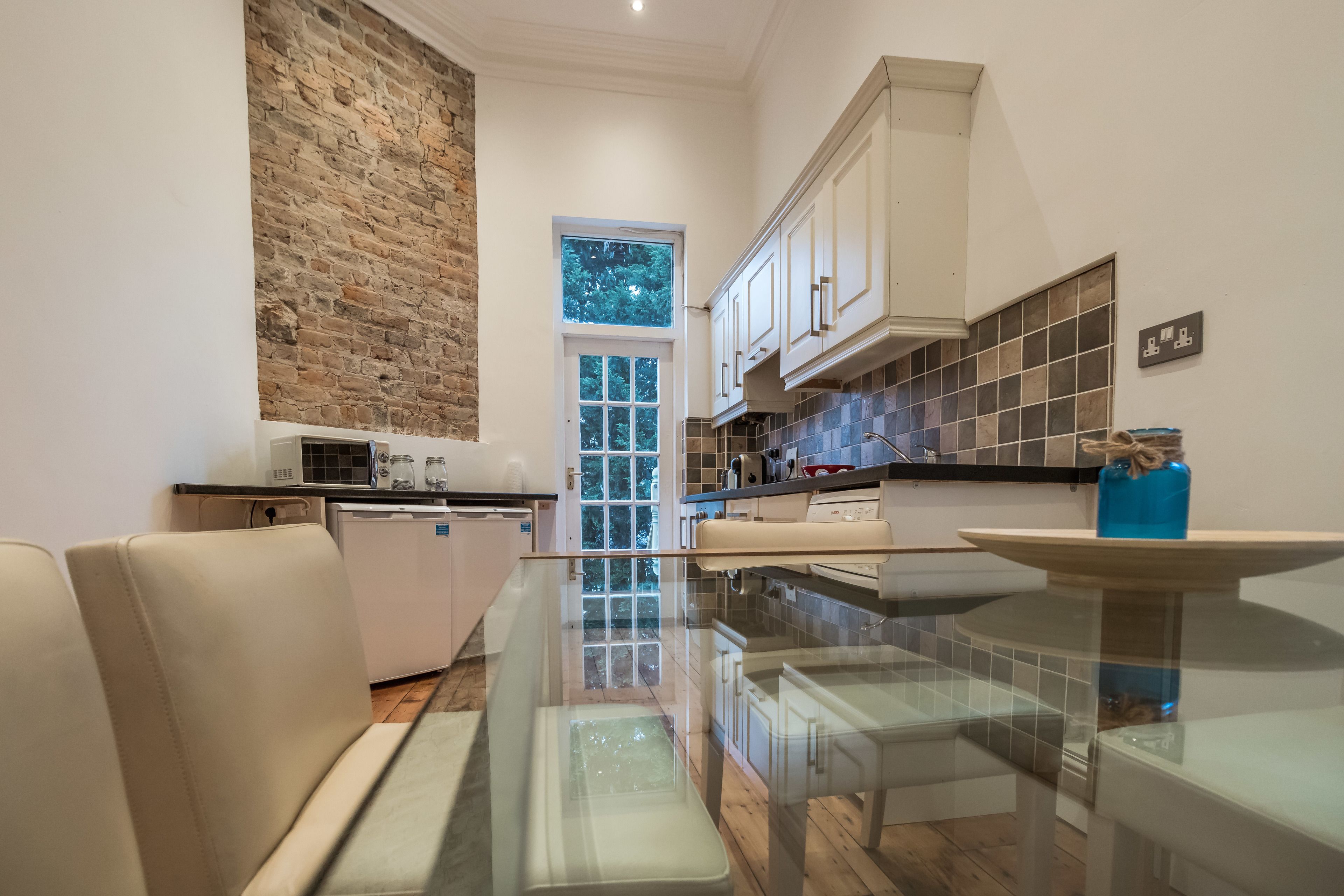 Une cuisine moderne avec une table à manger en verre, des armoires blanches et un mur d’accent en briques.