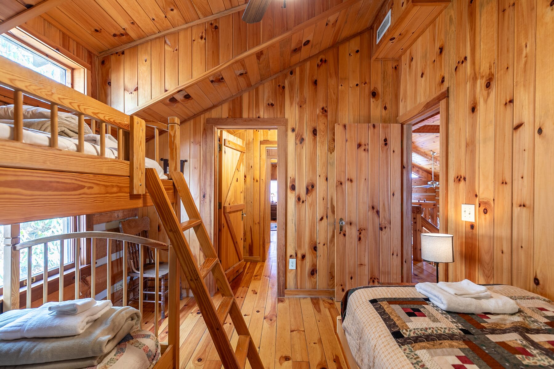L’intérieur d’un chalet en bois, avec un lit superposé, une échelle, un lit recouvert d’une courtepointe et une porte donnant sur une autre pièce.