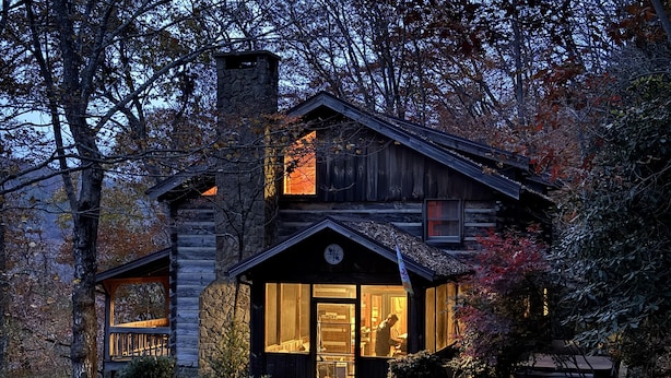 Une cabane rustique en rondins de bois, dotée d’une cheminée en pierre, de grandes fenêtres et d’une entrée bien éclairée.