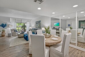Sala de estar moderna com uma mesa de jantar redonda, cadeiras brancas e um espelho grande que reflete o ambiente.