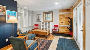 A cozy living room with a wooden coffee table, two blue armchairs, a red chair, a lamp, and a wall with wine bottle labels.