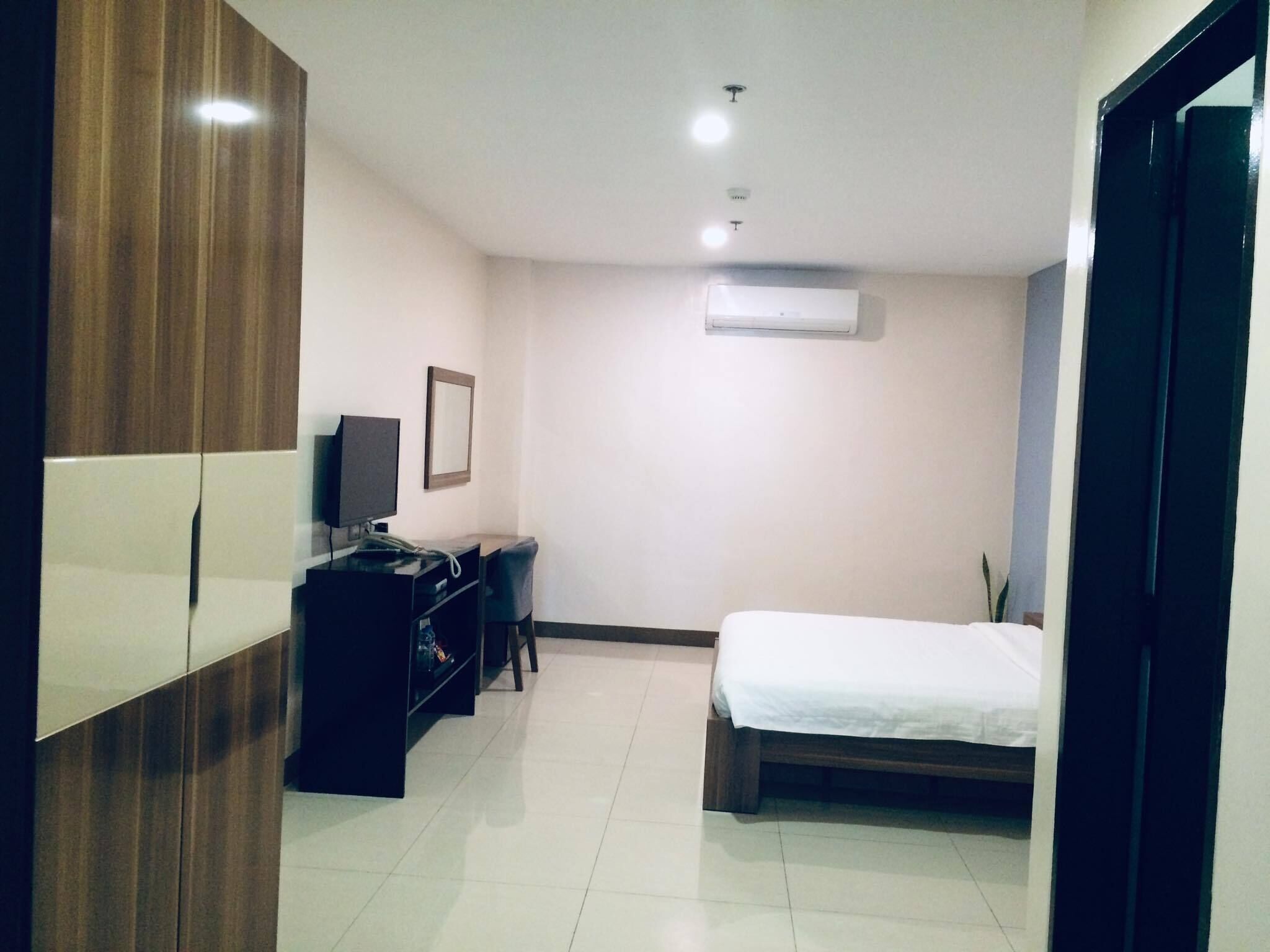 A hotel room with a bed, a desk with a computer, a chair, a wardrobe, and an air conditioning unit.