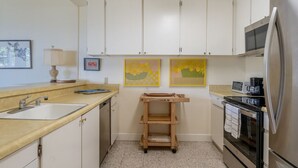 Cocina compacta con armarios blancos, un lavamanos, microondas y refrigerador. Hay dos obras de arte enmarcadas en la pared y un carrito de madera en el centro.