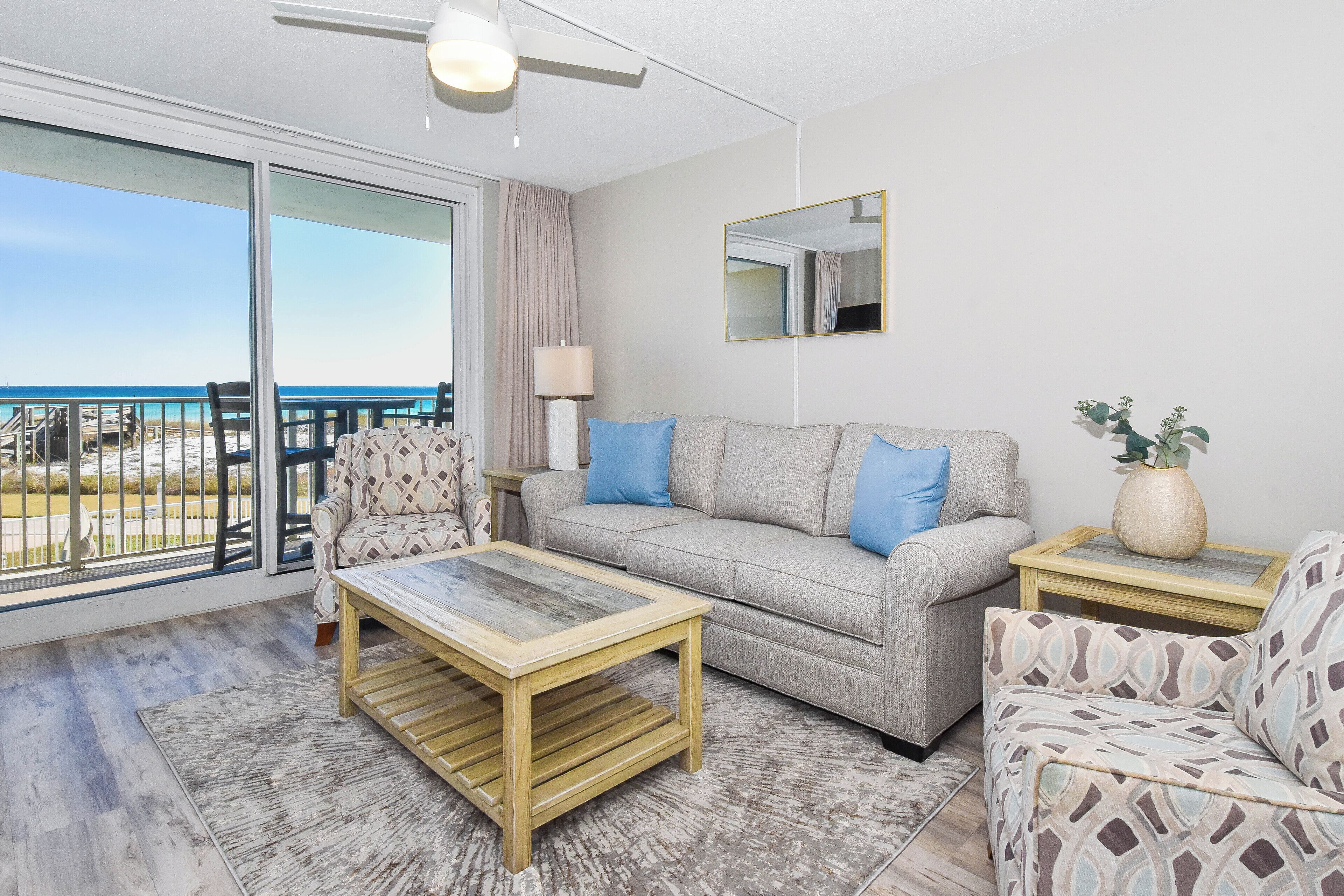 A living room with a sofa, a coffee table, a patterned armchair, a lamp, a mirror, and a view of the beach through large windows.