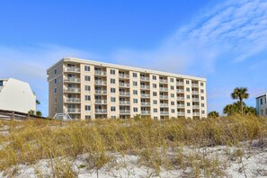 A multi-story hotel with balconies, situated on a sandy area with sparse vegetation. - Coastal decor w/a balcony & beachfront, pool view! 🌅 (Destin)