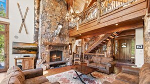 A rustic interior with a stone fireplace, leather sofas, a wooden staircase, and a large chandelier. - Bullshead By Tahoe Getaways - 4700SQ FT Luxury Riverfront Estate w Hot Tub (Olympic Valley)