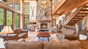 A log cabin interior with a stone fireplace, leather sofas, and a wooden staircase. - Bullshead By Tahoe Getaways - 4700SQ FT Luxury Riverfront Estate w Hot Tub (Olympic Valley)
