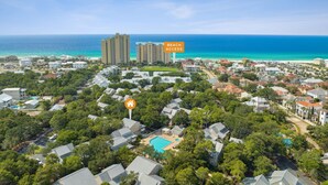 A residential area with a large swimming pool surrounded by houses, a beach access sign, and high-rise buildings in the background. - Family-Friendly 4BR Cottage with Pool View - Hidden Dunes Resort (Miramar Beach)