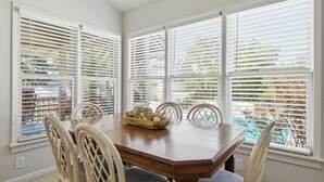 A dining area with a wooden table and chairs, large windows with blinds, and a view of a pool and greenery outside. - Family-Friendly 4BR Cottage with Pool View - Hidden Dunes Resort (Miramar Beach)