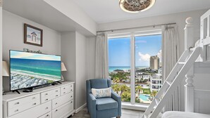 A room with a TV, a chair, a bed, a staircase, and a large window with a view of the ocean and buildings. - Gulf Front ~ Private Balcony ~ Pool (Miramar Beach)