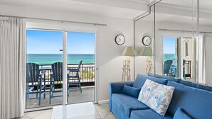 A room with a blue sofa, a view of the ocean, a sliding glass door, and two wall clocks. - Gulf Front ~ Private Balcony ~ Pool (Miramar Beach)