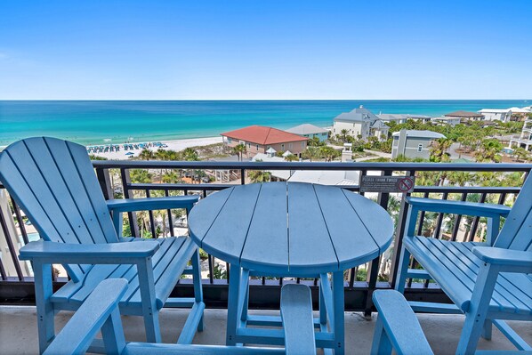 Un balcon avec des fauteuils Adirondack bleus et une petite table ronde, donnant sur une plage et les bâtiments d’un complexe hôtelier.