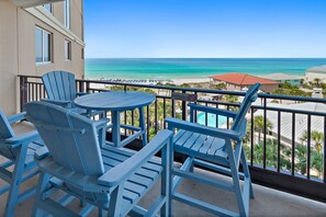 Un balcon avec des fauteuils Adirondack bleus et une table, donnant sur une plage parsemée de parasols et une piscine.